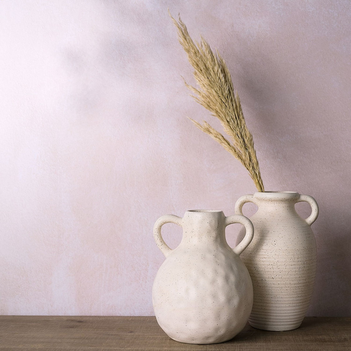 Round dimpled speckled ceramic vase in cream with two looped handles, shown beside a taller ribbed vase holding dried pampas grass against a pink backdrop.