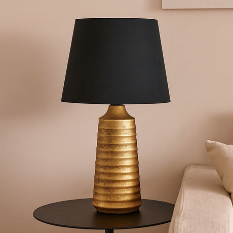 Ribbed ceramic table lamp with a ridged antique gold base and tapered black fabric shade, shown on a dark side table in a living room setting.