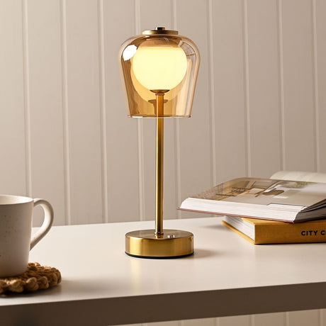 Hestia USB LED touch table lamp with bell-shaped amber glass shade, glowing white globe, slim gold stem and round gold base on a desk beside books and a mug.