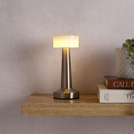 Tapered LED touch table lamp with round white shade and brushed silver stem, lit on a wooden shelf beside books and a plant.