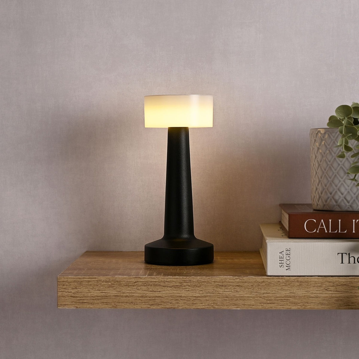 Matt black touch table lamp with a round white drum shade lit on a slender tapered stem, shown on a wooden shelf beside books and a plant.