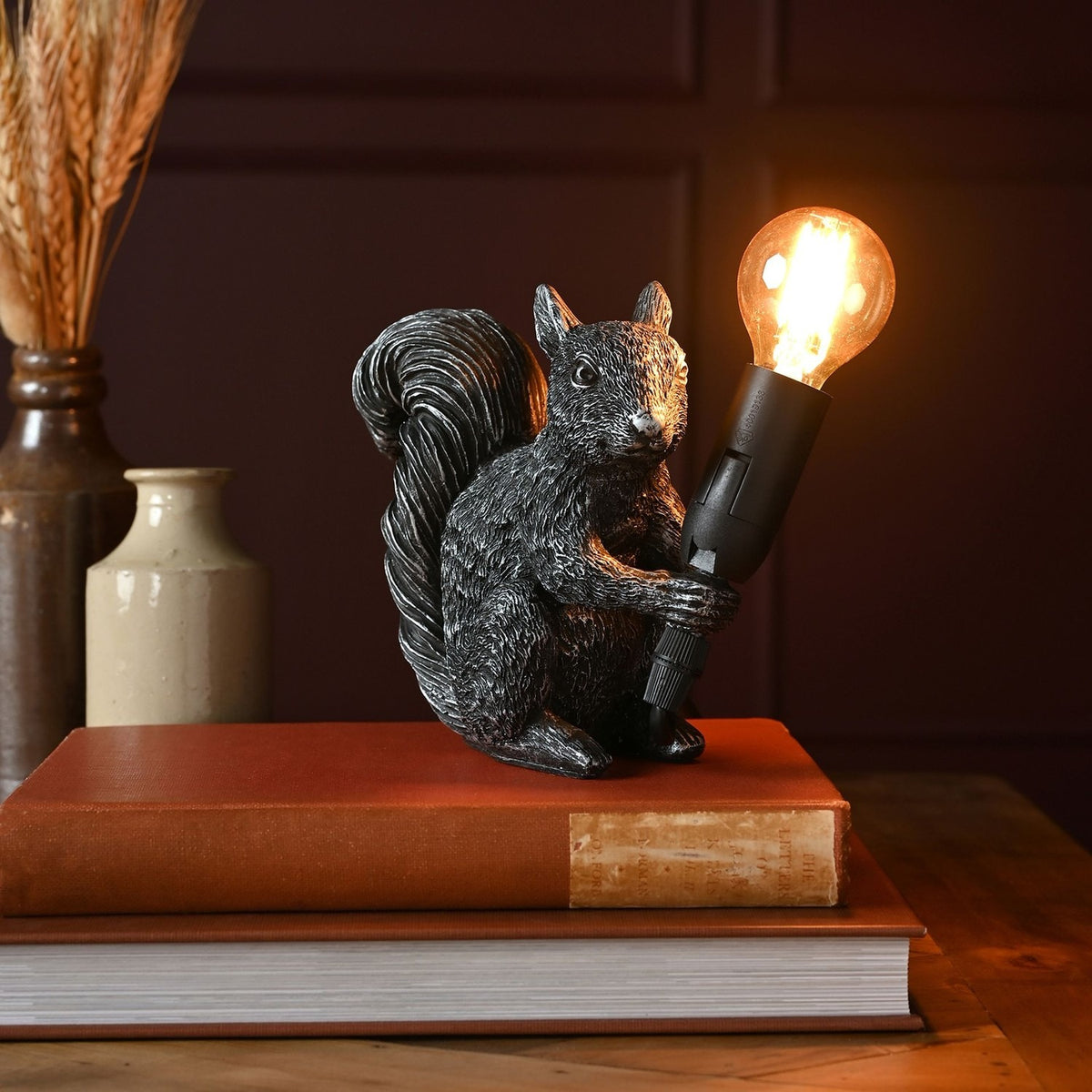 Squirrel Resin Table Lamp, 16cm, black finish with silvery highlights, holding an exposed bulb holder with glowing globe bulb, shown atop books on a wooden table.