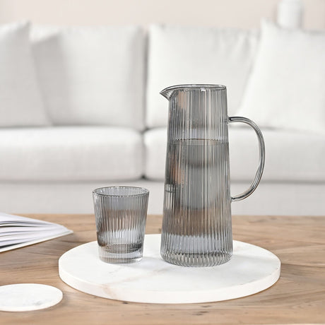 Tall Tapered Ribbed Jug with Curved Handle 1300ml in smoky grey glass, shown filled with water alongside a matching ribbed tumbler on a marble tray in a living room setting.