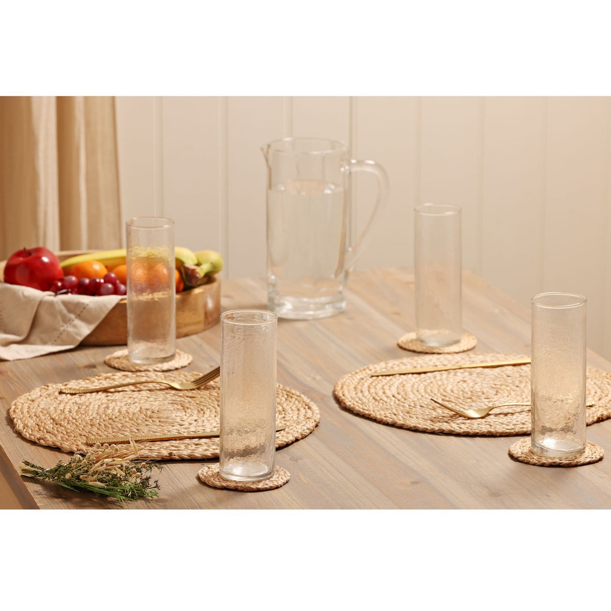Tall straight-sided hammered style clear drinking glasses on a dining table with a glass jug, woven placemats, gold cutlery, and fresh fruit.