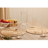 Tall straight-sided hammered style clear drinking glasses on a dining table with a glass jug, woven placemats, gold cutlery, and fresh fruit.