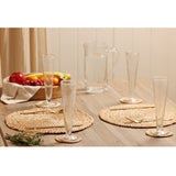 Conical footed hammered style glass champagne flutes on woven placemats with a water jug and fruit bowl on a wooden dining table.