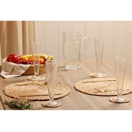 Conical footed hammered style glass champagne flutes on woven placemats with a water jug and fruit bowl on a wooden dining table.