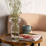 Clear ribbed glass vase with a rounded top and narrow waist, holding gypsophila stems on a wooden side table beside a mug and book.