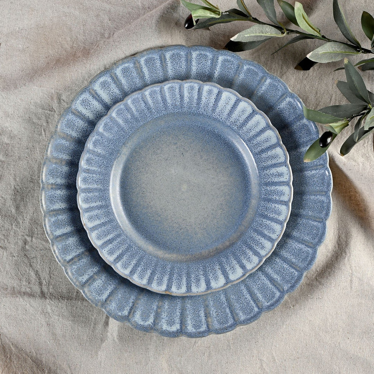 Small round fluted stoneware plate with scalloped edge in speckled blue glaze, shown stacked on a matching larger plate on a linen surface with olive leaves nearby.