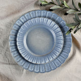 Small round fluted stoneware plate with scalloped edge in speckled blue glaze, shown stacked on a matching larger plate on a linen surface with olive leaves nearby.