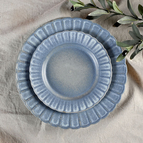 Small round fluted stoneware plate with scalloped edge in speckled blue glaze, shown stacked on a matching larger plate on a linen surface with olive leaves nearby.