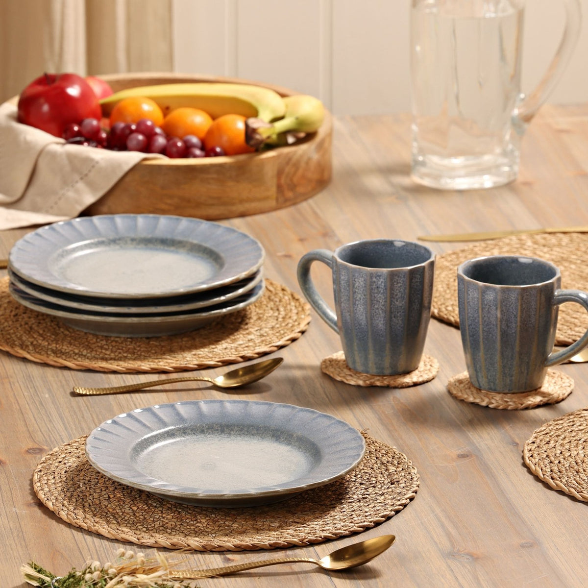 Small round fluted stoneware plates with speckled blue glaze stacked on a wicker placemat, styled on a wood table with mugs, fruit bowl, and cutlery.