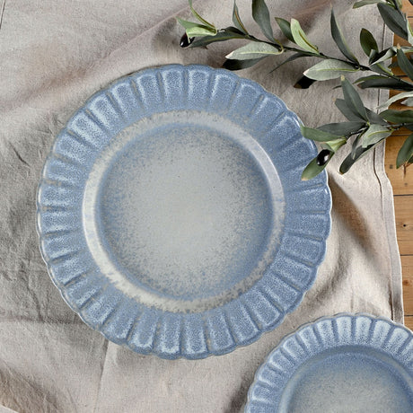 Round stoneware plate with a scalloped, fluted rim in mottled blue glaze fading to a pale grey-white centre, shown on linen with olive leaves.