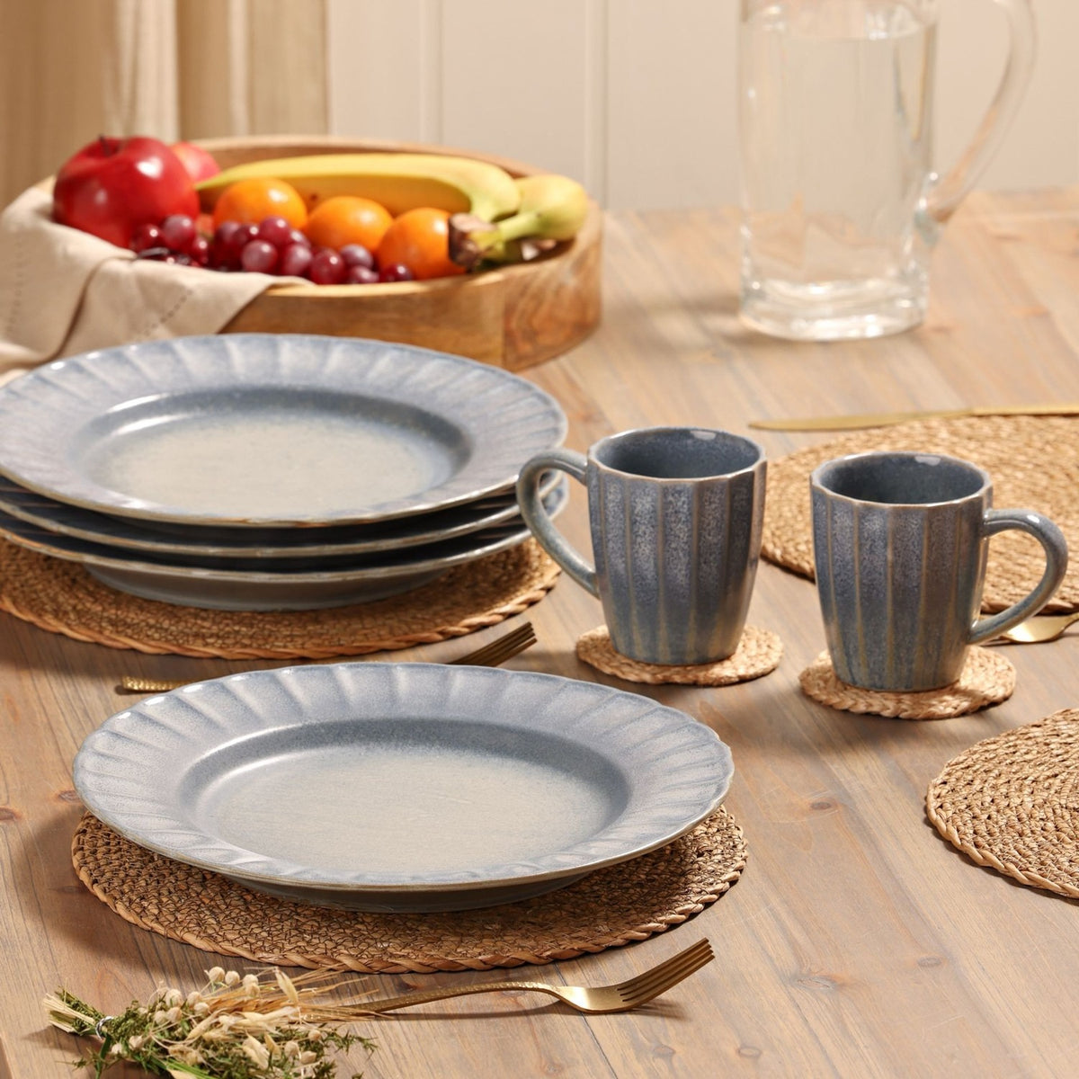 Round scalloped rim stoneware plates with mottled blue glaze, shown stacked and set on a wooden table alongside matching mugs, cutlery, and woven placemats.