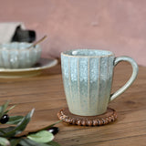 Tapered fluted stoneware mug with a speckled green glaze, lighter near the rim and deepening to grey-green at the base, shown with a ceramic handle on a wooden table.