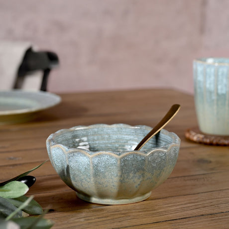 Scalloped fluted stoneware bowl in speckled green glaze with cream-edged ridges, shown on a wooden table with a spoon inside.