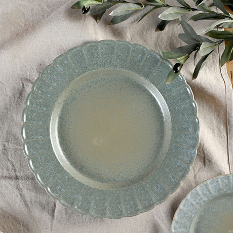 Round stoneware plate with a scalloped, fluted rim in speckled green glaze, deeper tone on the ridges and paler centre, shown on a neutral linen cloth with olive leaves.