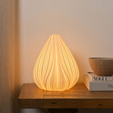 Ribbed Flower Bud ceramic table lamp in white, lit on a wooden console beside books and a bowl, showing its tapered petal-like shape and soft glow.