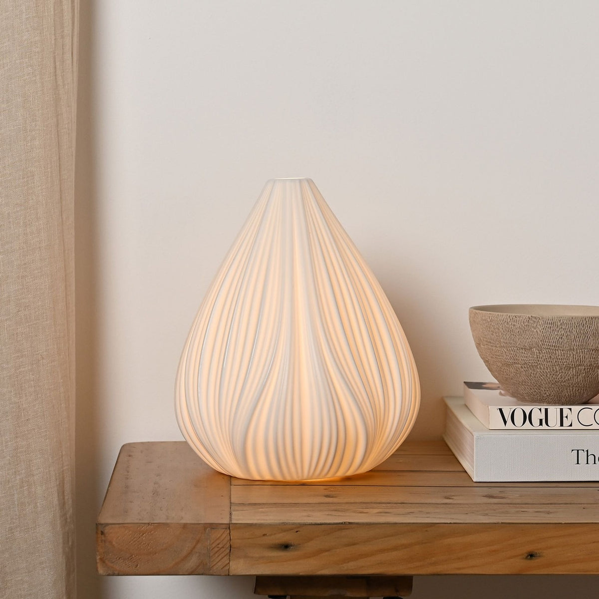 Ribbed Flower Bud ceramic table lamp in white, lit on a wooden console table beside a stack of books and a ceramic bowl.