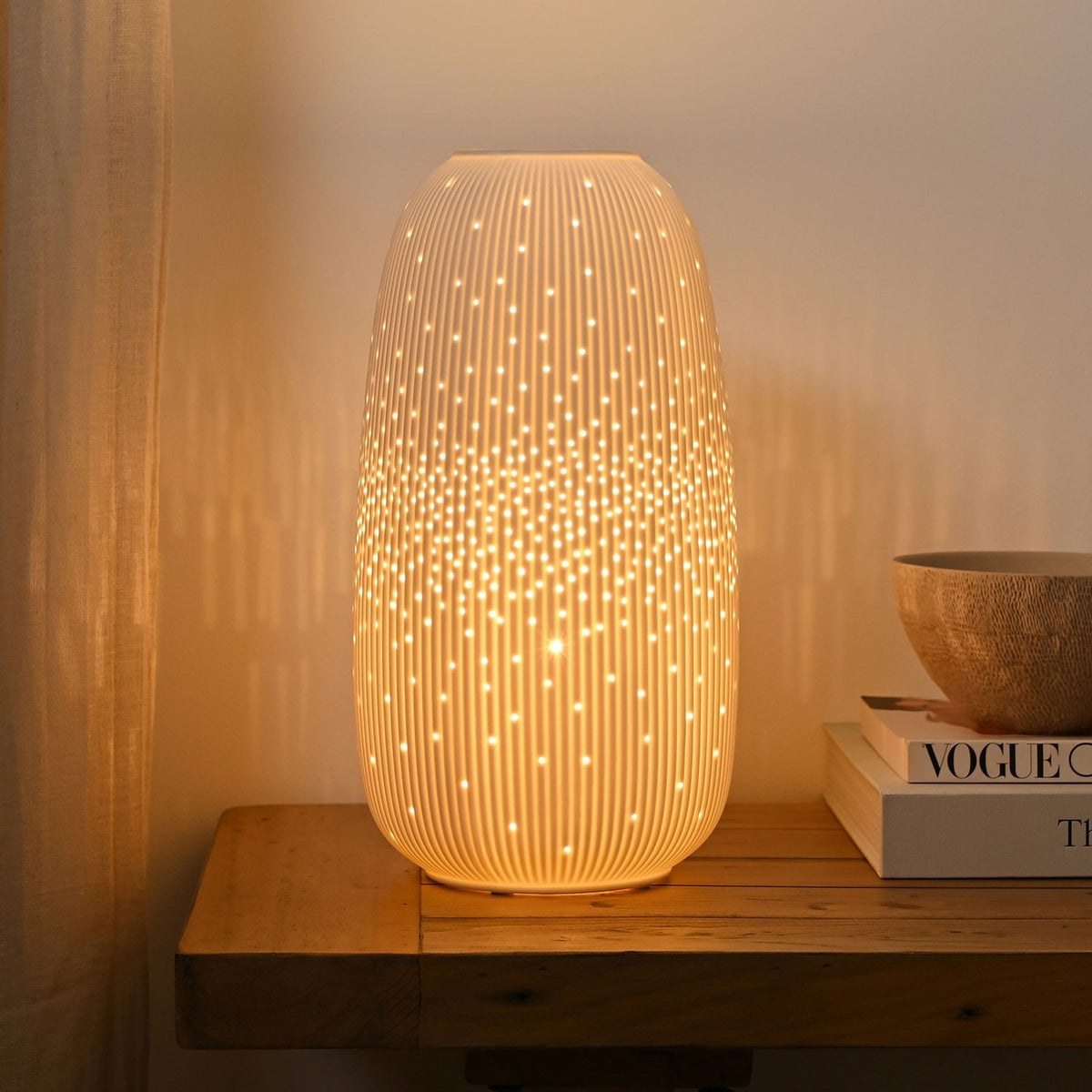 Tall Oval Ribbed Pierced Dot Ceramic Table Lamp, 33cm, in white, lit on a wooden console beside books and a bowl, glowing through fine vertical ribs and scattered pierced dots.