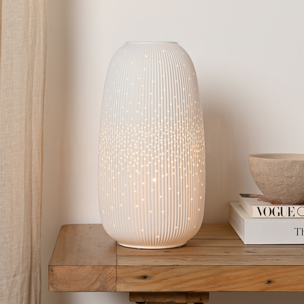 Tall Oval Ribbed Pierced Dot Ceramic Table Lamp 33cm in white, lit to show glowing dot pattern, displayed on a wooden console table beside books and a bowl.