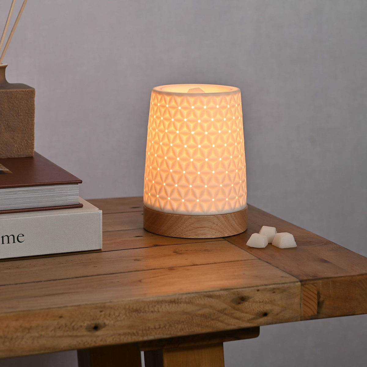 Tapered Embossed Star Aroma Ceramic Table Lamp in white with wood base, glowing on a wooden side table beside wax melts and stacked books.