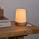 Tapered Embossed Star Aroma Ceramic Table Lamp in white with wood base, glowing on a wooden side table beside wax melts and stacked books.