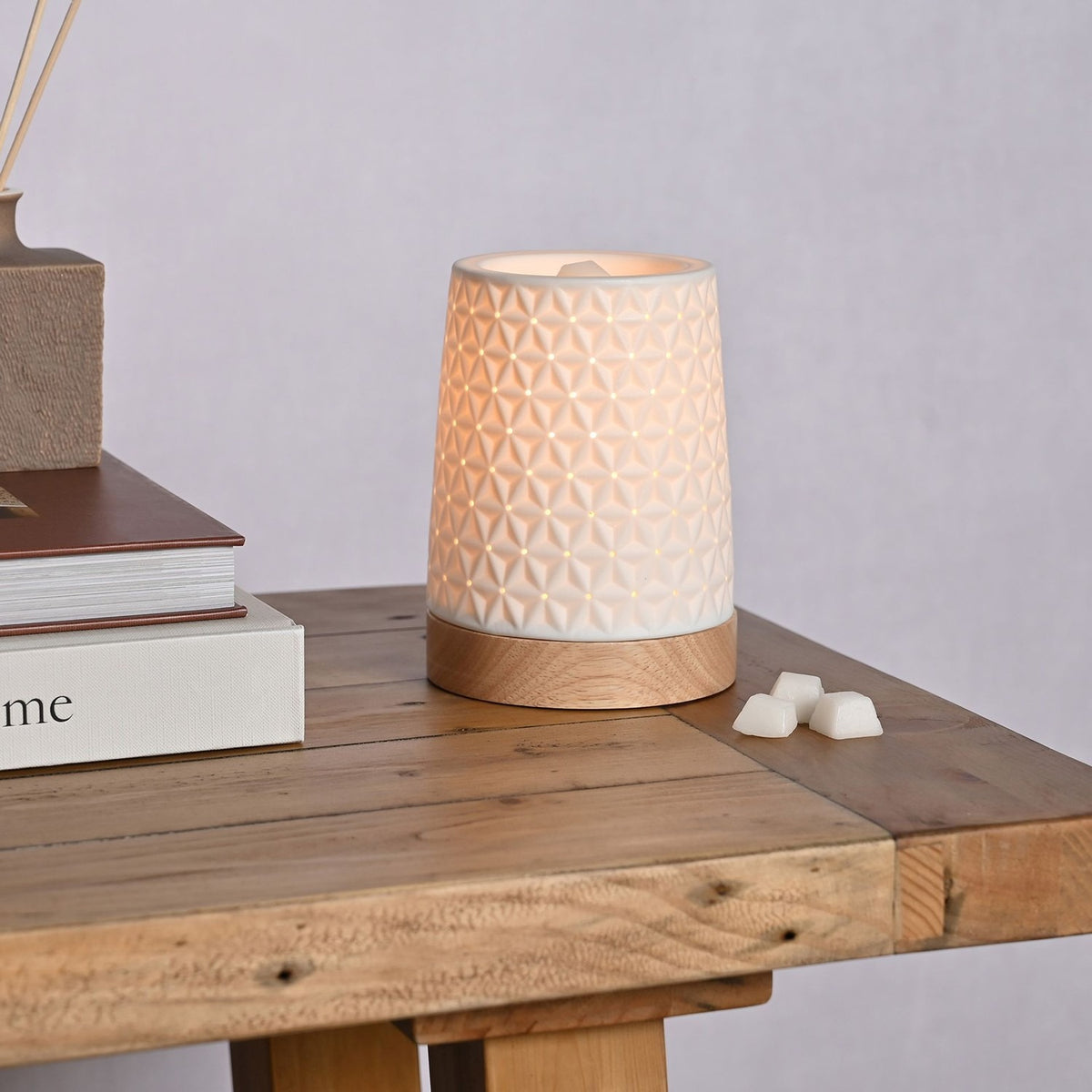 Tapered Embossed Star Aroma Ceramic Table Lamp, 15cm, in white with a star-patterned perforated design, on a round wooden base, shown lit on a wooden table beside wax melts and books.