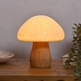Pierced Mushroom battery powered ceramic table lamp with a white pierced dome glowing with pinpoint light, set on a curved wood-tone stem on a wooden surface.