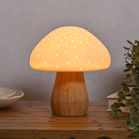 Pierced Mushroom battery powered ceramic table lamp with a white pierced dome glowing with pinpoint light, set on a curved wood-tone stem on a wooden surface.
