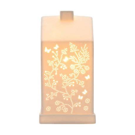 White ceramic table lamp shaped like a house with pitched roof and chimney, cut-out butterfly and floral vine design glowing warm white, on plain background.
