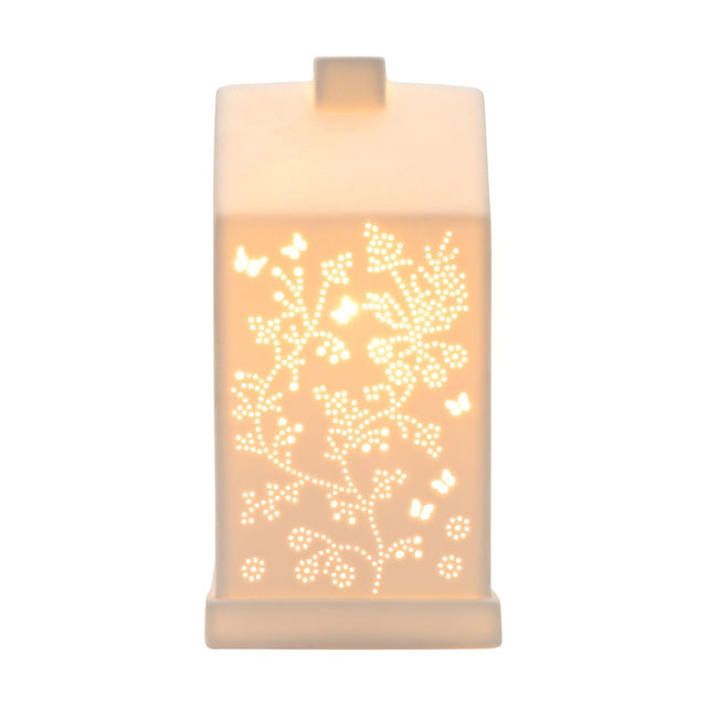 White ceramic table lamp shaped like a house with pitched roof and chimney, cut-out butterfly and floral vine design glowing warm white, on plain background.