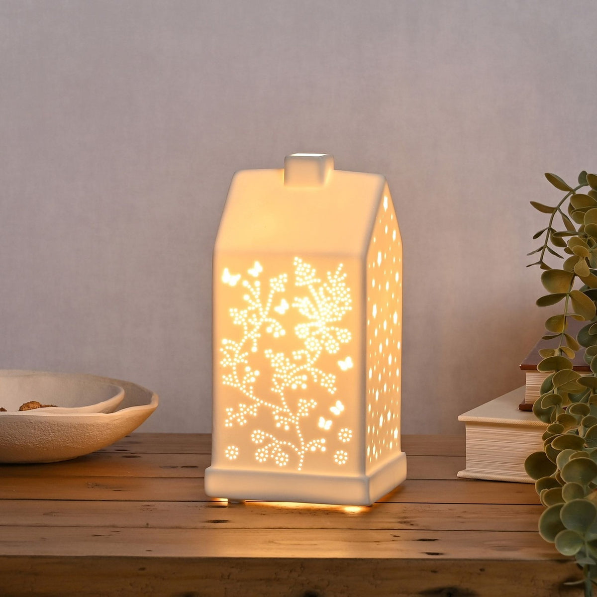 White ceramic table lamp shaped like a house with pitched roof and chimney, cut-out butterflies and floral vines glowing warmly, shown on a wooden table.
