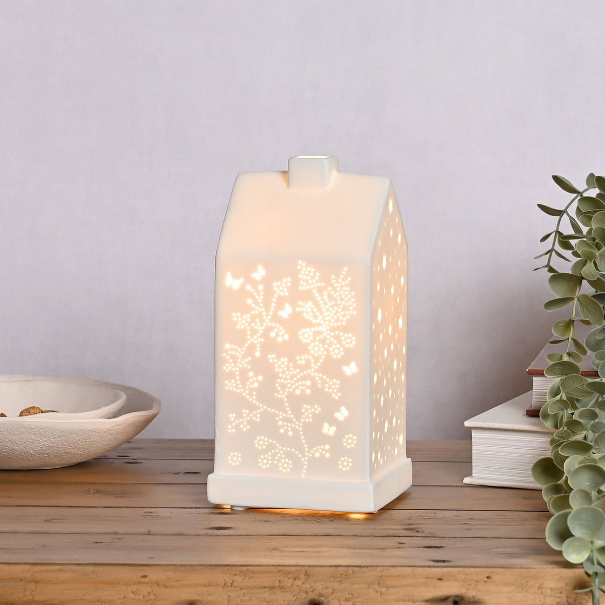 White ceramic house-shaped table lamp, 20.5cm, with cut-out butterflies and floral stems lit from within, shown on a wooden shelf beside books and greenery.