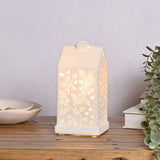White ceramic house-shaped table lamp, 20.5cm, with cut-out butterflies and floral stems lit from within, shown on a wooden shelf beside books and greenery.