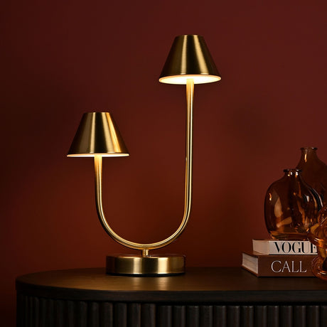 Curved Stem USB Touch Double Light Table Lamp in bronze, with a round base, curving U-shaped stem, and two cone shades at uneven heights, shown on a sideboard against a dark red wall.