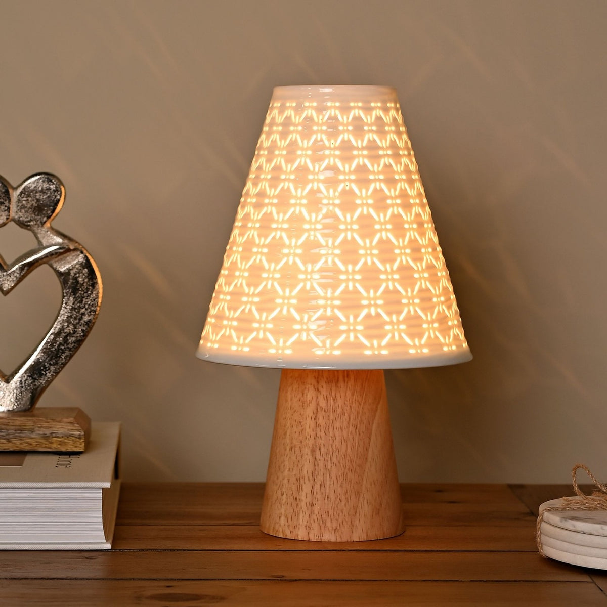 Pierced Cone Ceramic Table Lamp with wooden base, lit on a wooden sideboard beside books and a silver heart ornament, showing its white perforated geometric shade glowing warmly.