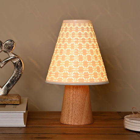 Pierced Cone Ceramic Table Lamp with wooden base, lit on a wooden sideboard beside books and a silver heart ornament, showing its white perforated geometric shade glowing warmly.