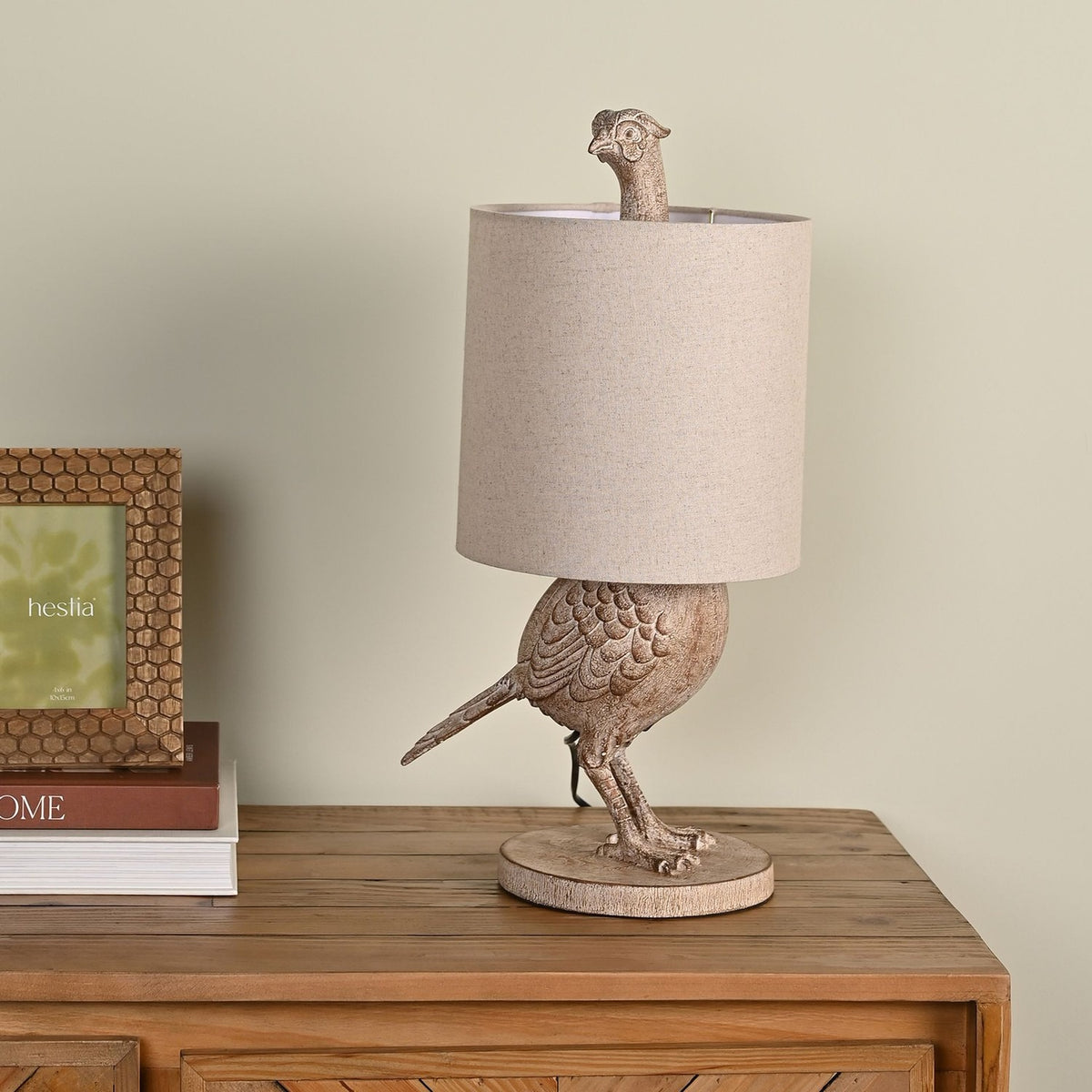 Wood effect pheasant table lamp, 54cm, with a beige drum shade the bird's head peeks above, light brown finish, shown on a wooden sideboard.
