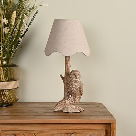 Owl Wood Effect Resin Table Lamp with scalloped beige shade, shown on a wooden sideboard beside dried florals in a neutral-toned room.