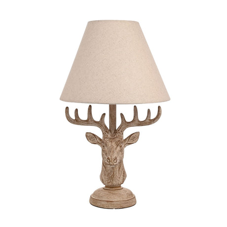 Table lamp with a wood effect stag's head base featuring textured fur, antlers and a round stepped plinth, topped with a beige conical shade, on a plain white background.