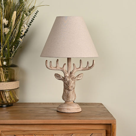 Wood effect stag's head table lamp with a beige fabric shade, textured antler and fur detailing on a round stepped base, shown on a wooden sideboard.