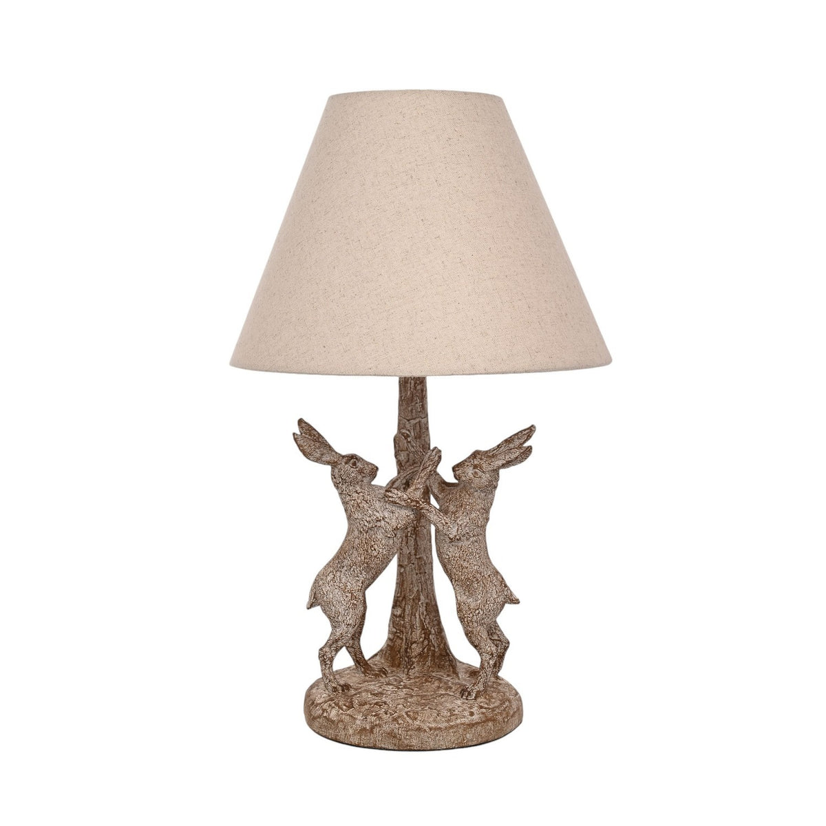 Table lamp with a wood-effect base of two hares facing each other beside a tree trunk, in brown with paler highlights, topped by a natural cone-shaped fabric shade, shown on a plain white background.