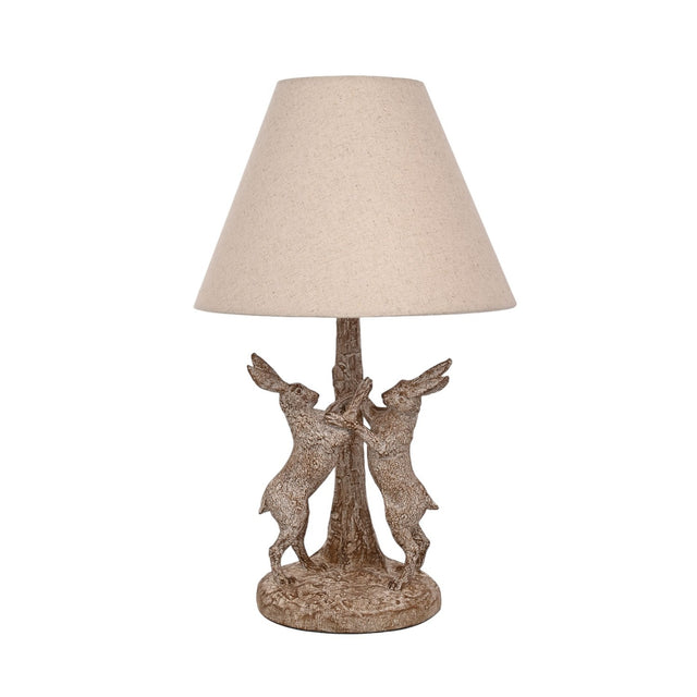 Table lamp with a wood-effect base of two hares facing each other beside a tree trunk, in brown with paler highlights, topped by a natural cone-shaped fabric shade, shown on a plain white background.