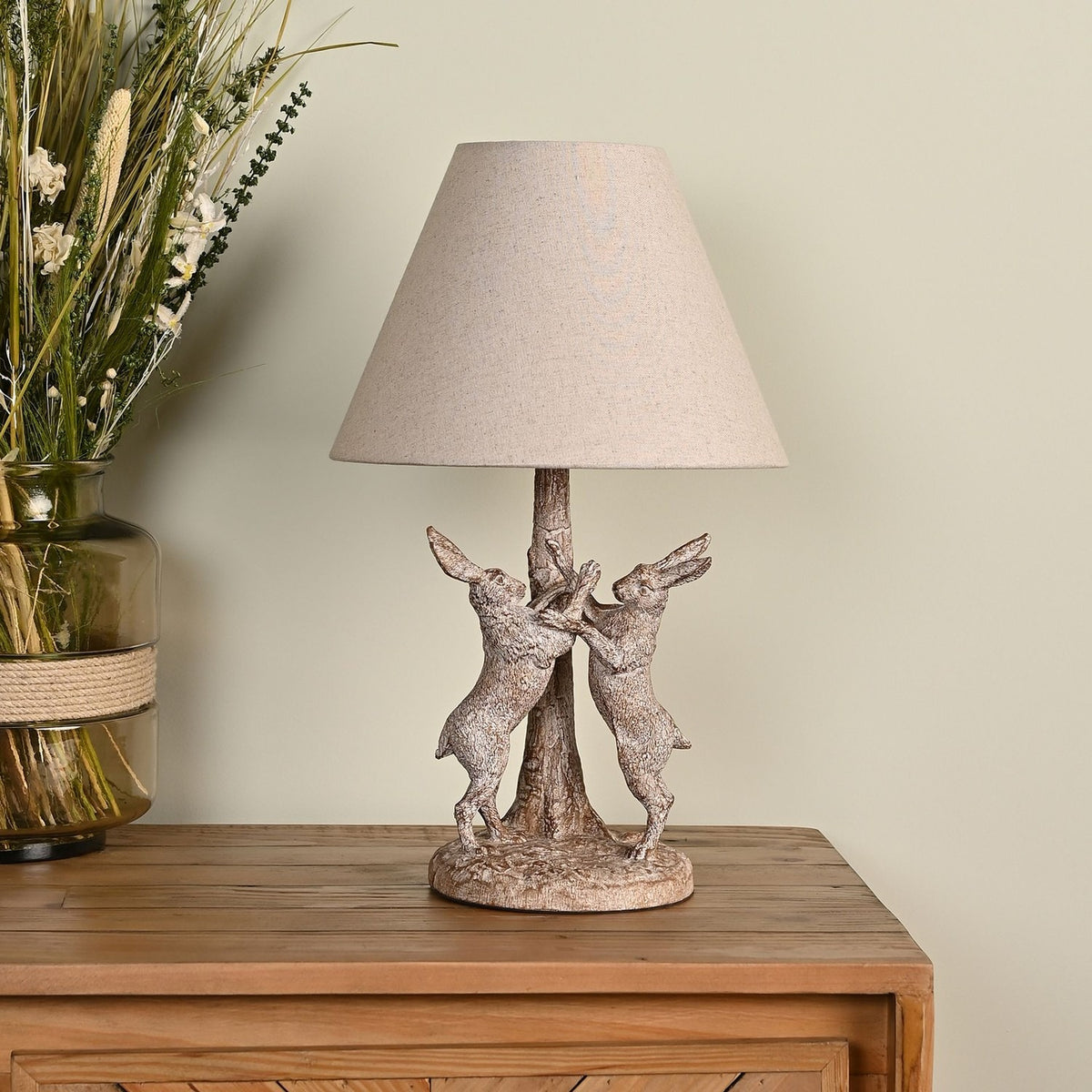 Wood Effect Hare Table Lamp with Natural Shade 46cm in Brown, featuring two boxing hares flanking a tree trunk base with a tapered fabric shade, shown on a wooden sideboard.