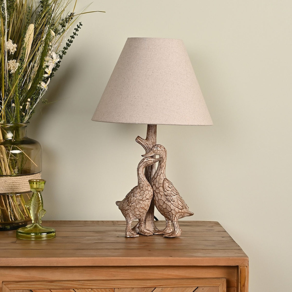 Table lamp with two sculpted geese on a tree-trunk stem in brown wood-effect resin, topped with a tapered beige fabric shade, shown on a wooden sideboard.