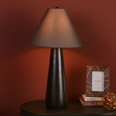 Tapered Resin Table Lamp, 69cm, with dark brown resin base and conical taupe fabric shade, shown lit on a side table against a dark red wall.