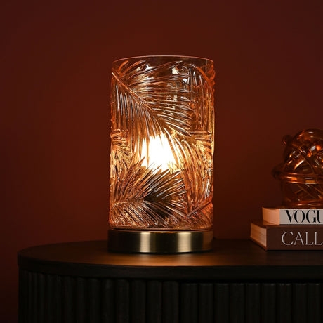 Palm Leaf Textured Glass Table Lamp, 23cm, with gold-toned base and cylindrical shade, lit on a dark cabinet beside books in a warm-toned room.