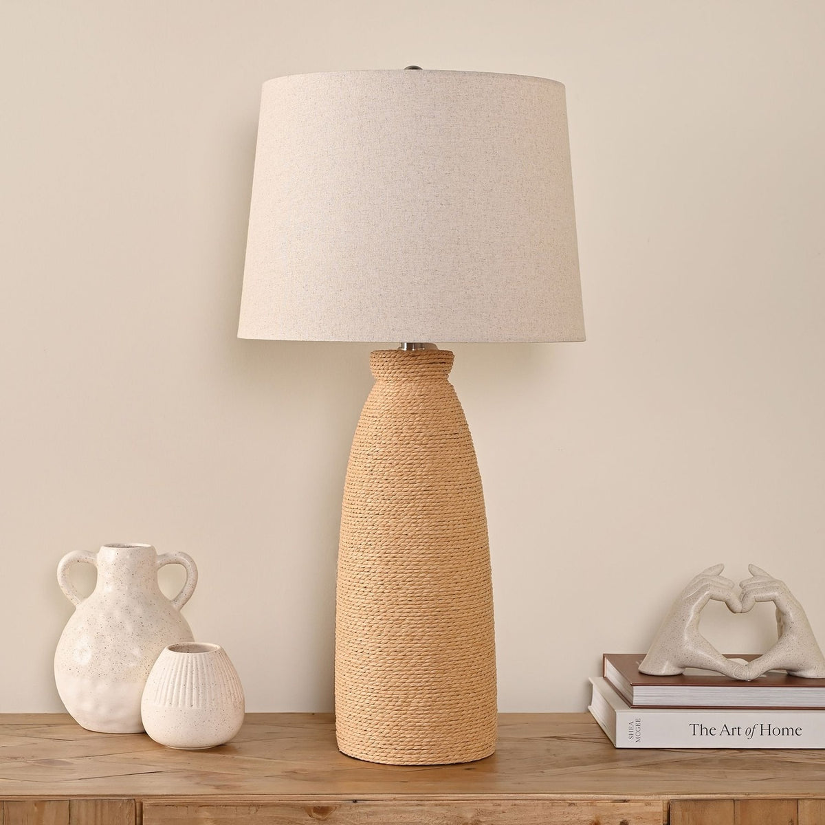 Tapered Rattan Table Lamp with Drum Shade 76cm in Natural, shown on a wooden console beside ceramic vases and stacked books in a living room setting.