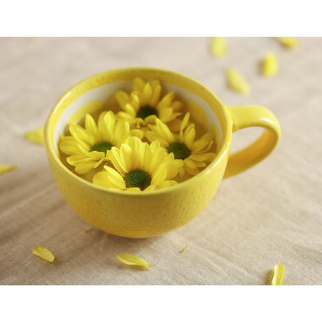 Round earthenware mug in glossy yellow with a lemon-inspired speckled finish and loop handle, shown filled with yellow daisy flowers on a neutral fabric surface.